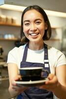 portrait de souriant asiatique femme dans tablier, barista donnant vous tasse de café, travail dans café, portion les boissons photo