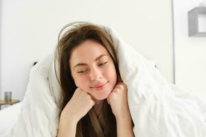 mignonne fille avec désordonné cheveux, mensonge dans lit couvert dans blanc feuilles couette, souriant et en riant coquette, dépenses temps dans sa chambre photo