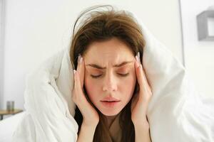 proche en haut de femme sentiment indisposé à maison, mensonge dans lit en dessous de blanc couvertures et renfrogné, émouvant diriger, a mal de tête, migraine, récupérer de convoitise photo