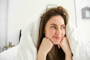 mignonne fille avec désordonné cheveux, mensonge dans lit couvert dans blanc feuilles couette, souriant et en riant coquette, dépenses temps dans sa chambre photo