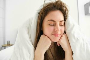 de bonne humeur femme sentiment confortable dans lit, mensonge dans chambre couvert dans blanc feuilles, souriant heureux, a désordonné cheveux dans matin, veille en haut et à la recherche content photo