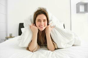 mignonne fille avec désordonné cheveux, mensonge dans lit couvert dans blanc feuilles couette, souriant et en riant coquette, dépenses temps dans sa chambre photo