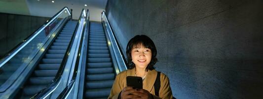 mignonne Jeune femme Aller vers le bas le escalier mécanique à le tube, en utilisant métro métro à commuer à travail ou université, permanent avec téléphone intelligent photo