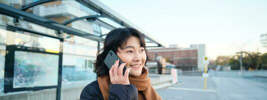 sourire coréen fille parlant sur mobile téléphone, permanent sur autobus arrêt, en utilisant téléphone intelligent, posant sur route dans hiver, enveloppé dans foulard, portant noir veste photo