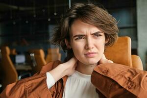 portrait de femme se sent douleur dans cou, est assis dans Bureau avec ordinateur portable, a tension dans muscles après travail tout journée photo