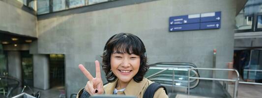 mignonne et élégant coréen fille, porte écouteurs, prend selfie sur téléphone intelligent, touristique enregistrements vidéo ou fait du une photo, des stands sur rue photo