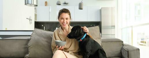 Matin et mode de vie concept. content Jeune femme sur canapé, séance avec chien, caresse sa chiot et en mangeant céréales, ayant petit-déjeuner, en riant et souriant photo