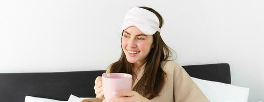 content Jeune femme départ sa Matin avec tasse de café dans lit, souriant et profiter sa paresseux jour, a en train de dormir masque sur front photo