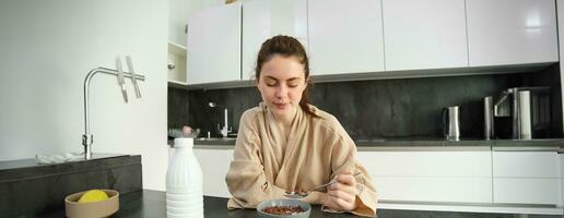 image de content Jeune femme en mangeant céréales à Accueil avec lait, ayant sa petit-déjeuner, portant peignoir de bain, séance dans cuisine seul photo