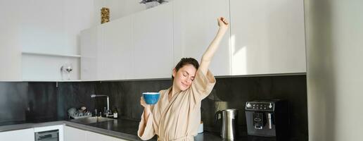 portrait de content fille dansant avec café dans le cuisine, portant peignoir de bain, profiter sa Matin routine photo