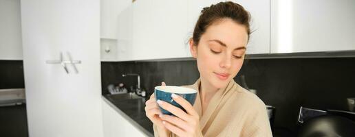 portrait de content, souriant moderne femme, départs sa journée avec Matin agresser de thé. fille en buvant café dans le cuisine, permanent avec agresser et à la recherche détendu photo