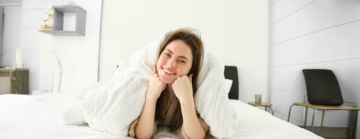 portrait de magnifique souriant femme, mensonge dans lit couvert dans couette, a désordonné cheveux, à la recherche content, relaxant dans sa chambre à coucher, dépenses temps dans Hôtel pièce dans Matin photo