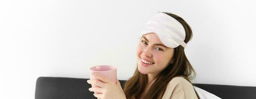 content Jeune femme départ sa Matin avec tasse de café dans lit, souriant et profiter sa paresseux jour, a en train de dormir masque sur front photo