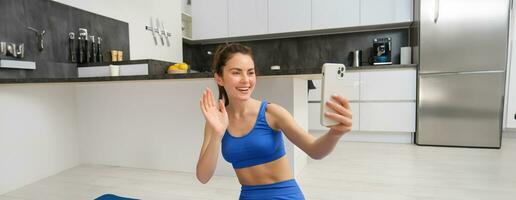 portrait de Jeune aptitude fille, blogueur enregistrements sa faire des exercices de maison, pose pour selfie sur téléphone intelligent, Est-ce que yoga sur caméra, est assis dans bleu soutien-gorge de sport et leggings photo