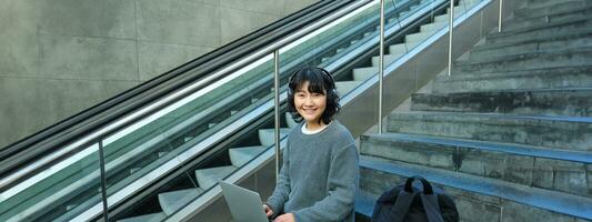 portrait de magnifique Jeune coréen femme, étudiant est assis sur escaliers dans Publique lieu, écoute la musique dans écouteurs, travaux sur projet sur ordinateur portable, travaux à distance photo