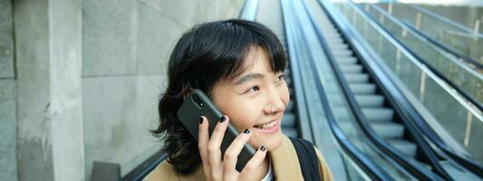 photo du visage de souriant coréen femme avec téléphone intelligent, fait du une téléphone appel, va vers le bas escalier mécanique dans ville, les déplacements à Université