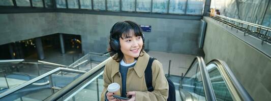 portrait de souriant coréen fille les boissons café à aller, va en haut un escalier mécanique, détient téléphone intelligent, visites Nouveau ville, arrive à train gare, écoute la musique dans écouteurs photo