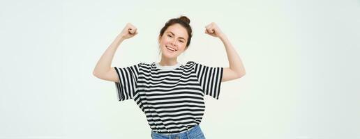 femme Puissance et féminisme. Jeune fille sentiment habilité et fort, fléchissement sa biceps, montrant muscles sur bras, permanent plus de blanc Contexte photo