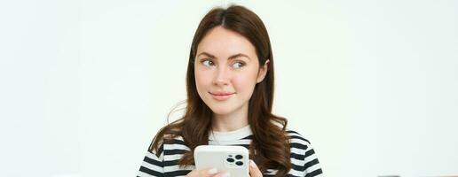 portrait de fille avec téléphone, femme en portant téléphone intelligent et pensée, souriant et à la recherche de côté, décider à acheter qch en ligne, blanc Contexte photo