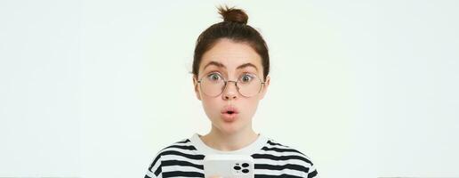 image de fille dans lunettes, en portant téléphone intelligent et à la recherche surpris, étonné par qch sur mobile téléphone, des stands plus de blanc Contexte photo