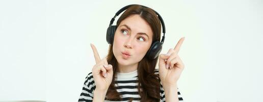 portrait de Jeune brunette femme dans écouteurs, écoute la musique dans écouteurs, montrer du doigt les doigts en haut, montrant bannière, publicité, isolé plus de blanc Contexte photo