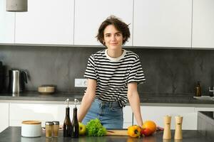 portrait de magnifique Jeune femme, femelle modèle posant près couper planche avec des légumes et salade pansement, fabrication se lumière régime repas, végétalien nourriture pour petit-déjeuner, permanent dans cuisine photo