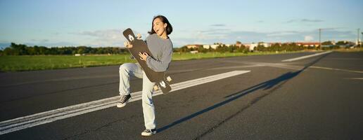 magnifique asiatique adolescent fille en jouant avec sa longboard, en portant planche à roulette comme si en jouant guitare, permanent sur route sur ensoleillé journée photo