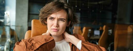 portrait de femme se sent douleur dans cou, est assis dans Bureau avec ordinateur portable, a tension dans muscles après travail tout journée photo