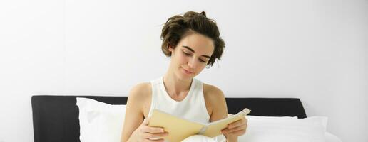 proche en haut portrait de Jeune femme, en train de lire journal, séance dans lit et en étudiant, réviser pour examen avant Aller à sommeil photo