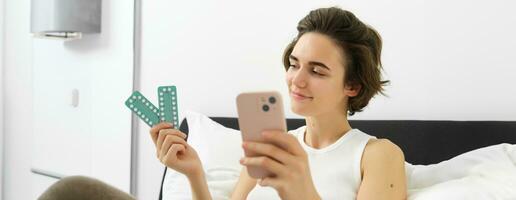 portrait de femme mensonge dans lit avec médicament, en portant pilules et téléphone intelligent, recherche pour côté effets ou dosage, en train de lire instruction avant prise comprimés photo