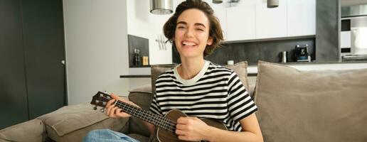 portrait de Jeune moderne femme, musicien en jouant ukulélé à maison, cueillette accords pour Nouveau chanson, séance sur canapé dans vivant pièce et souriant photo