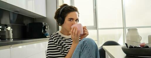 proche en haut portrait de souriant brunette femme, étudiant les boissons sa thé et écoute la musique ou ebook dans écouteurs, est assis dans cuisine et se détend photo