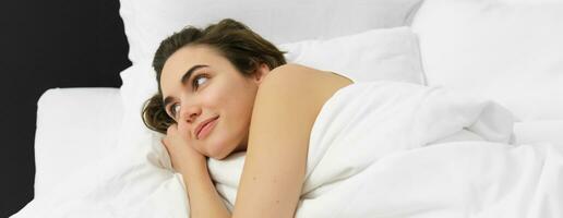 portrait de Jeune femme relaxant, mensonge dans lit sur doux blanc oreillers, couvert dans lin confortable feuilles, souriant et à la recherche à l'extérieur avec rêveur sourire photo