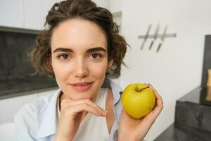 proche en haut portrait de Jeune femme en portant un pomme, en mangeant en bonne santé des fruits dans sa cuisine, souriant Heureusement photo