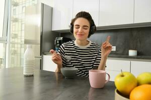portrait de content femme jouit écoute à la musique tandis que en mangeant céréales pour petit-déjeuner, portant sans fil écouteurs, souriant, séance dans le cuisine photo