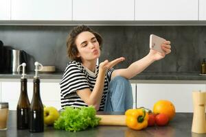 mignonne Jeune femme, mode de vie blogueur cuisson, séance dans le cuisine avec des légumes et couper conseil, prise selfie sur sa Nouveau téléphone intelligent, Envoi en cours baisers à mobile caméra photo