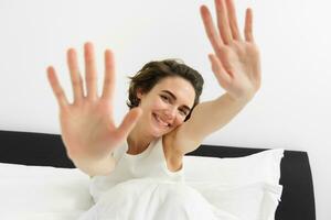 magnifique Jeune femme élongation sa mains vers caméra, veille en haut dans matin, repos dans sa lit, souriant et à la recherche content photo