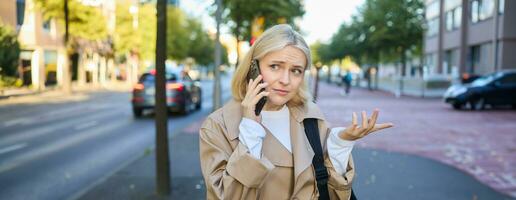 portrait de femme avec confus affronter, parlant sur mobile téléphone, répondre téléphone appel, permanent sur rue avec perplexe, Aucune idée expression, posant sur route photo