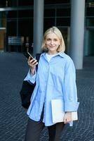 verticale portrait de Jeune Bureau directeur, femme dans bleu collier chemise et sac à dos, en portant portable et téléphone intelligent, attendre près affaires bâtiment sur rue, souriant à caméra photo