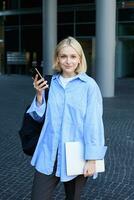 verticale portrait de Jeune Bureau directeur, femme dans bleu collier chemise et sac à dos, en portant portable et téléphone intelligent, attendre près affaires bâtiment sur rue, souriant à caméra photo