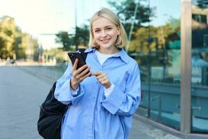 portrait de souriant, moderne blond femme en marchant le long de le rue dans bleu collier chemise, a sac à dos sur épaule, Messagerie, en utilisant mobile téléphone app photo