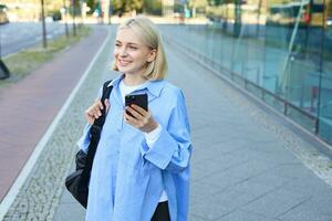 portrait de souriant, moderne blond femme en marchant le long de le rue dans bleu collier chemise, a sac à dos sur épaule, Messagerie, en utilisant mobile téléphone app photo