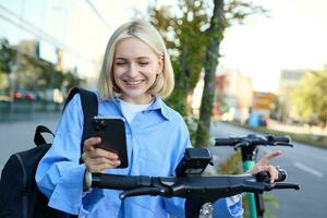 portrait de Jeune souriant femelle modèle, location un électrique scooter, en utilisant mobile téléphone à analyse qr code avec téléphone intelligent application, équitation Accueil de université, permanent en plein air sur rue photo