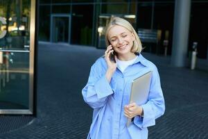 portrait de Jeune femme d'affaires dans bleu collier chemise, parlant sur mobile téléphone, permanent près Bureau bâtiment et souriant, en portant travail portable photo