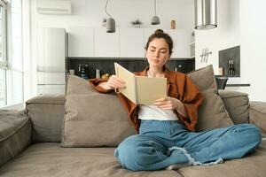 souriant Jeune brunette femme, est assis sur canapé dans vivant chambre, détient carnet de notes, lit sa Remarques, études pour examen, étudiant Est-ce que sa devoirs à Accueil photo