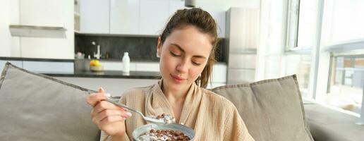 image de souriant, content Jeune femme en mangeant petit-déjeuner, en portant bol de céréales avec lait, ayant repas à Accueil photo