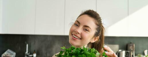 portrait de magnifique femme cuisson, en portant Frais persil, ajouter herbes tandis que fabrication salade, en bonne santé repas dans le cuisine photo