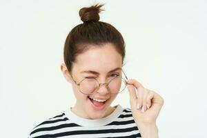 proche en haut portrait de élégant moderne fille, portant lunettes, souriant et un clin d'oeil à caméra, permanent plus de blanc Contexte photo