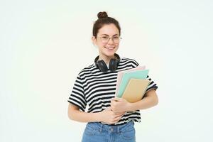 éducation concept. souriant brunette fille, étudiant dans décontractée vêtements, détient sa livres, étude matériel, porte écouteurs plus de cou, regards sur de soi et content, isolé plus de blanc Contexte photo