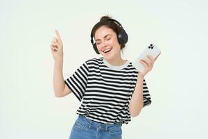 Jeune femme avec téléphone intelligent écoute à musique, dansant à sa préféré chanson dans écouteurs, posant contre blanc Contexte photo